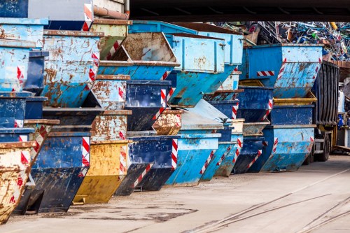 A row of large metal industrial skips lined up on a paved surface, primarily painted blue with some showing signs of rust and wear. The skips are filled with mixed waste materials, including crumpled paper, plastic, and other refuse, with open tops revealing the contents inside. The surrounding environment appears to be an outdoor waste management or rubbish collection area, possibly adjacent to a commercial or industrial site in Tunbridge Wells, as suggested by the context of local rubbish removal services. To the right, a black waste container or recycling bin is partially visible, and the background includes a dark structure or wall. The lighting suggests daytime with natural sunlight illuminating the scene. The image depicts typical rubbish disposal infrastructure used by companies like House Clearance Tunbridge Wells, emphasizing the handling and staging of waste for collection or disposal within a designated area.