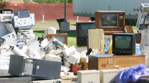 A large outdoor pile of discarded electronic waste and old appliances, including broken computer monitors, CRT televisions with wooden frames, various boxed and unboxed electronic devices, and tangled cables, situated on a paved surface next to a grassy area and a red wall. The items display a range of textures from the smooth, glossy screens and ceramic components of the monitors to the wooden casings of the older televisions. Some devices are stacked haphazardly, with parts spilling onto the ground, while others lean against each other. The scene suggests a collection of waste awaiting removal or disposal, fitting within the scope of rubbish removal services provided by House Clearance Tunbridge Wells. In the background, there is a glimpse of trees and a fence, with natural daylight illuminating the scene, emphasizing the cluttered nature of the waste and the variety of materials involved, typical of electronic waste collection in the local area around Tunbridge Wells.
