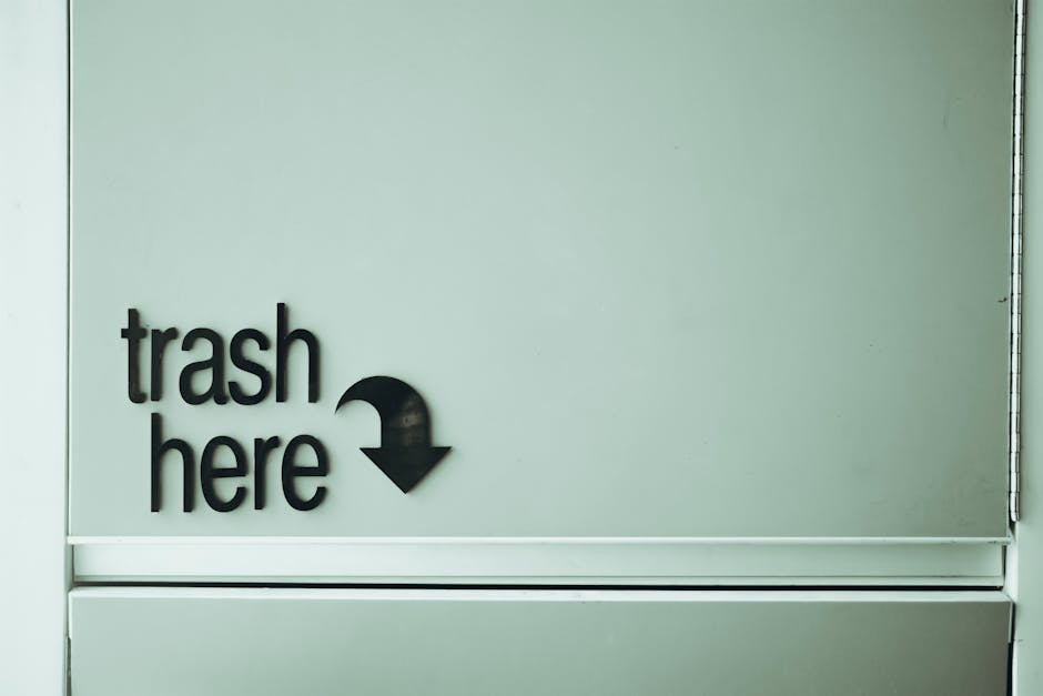 A collection of overflowing waste and rubbish bags, including black plastic bin liners, cardboard boxes, paper and packaging waste, and various miscellaneous items piled on a paved area near a metal r