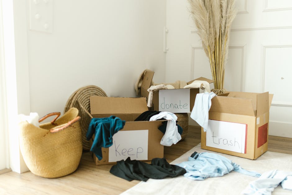 Indoor scene showing several cardboard boxes placed on a wooden floor near a white wall and a white door. One box is labeled 'Keep' and contains clothing items, including a blue shirt and black pieces