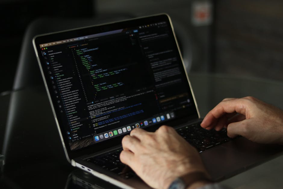 A modern silver laptop with a black keyboard sits open on a light wooden desk surface in an indoor setting, displaying lines of code or script on its bright screen. The laptop is positioned towards th
