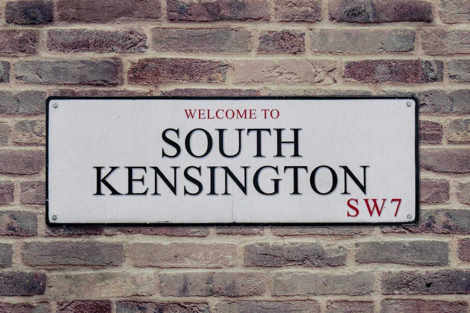 A rectangular metal street sign mounted on a red and brown brick wall, displaying the text 'Welcome to South Kensington' in black capital letters, with 'SW7' in red at the bottom right corner. The sig